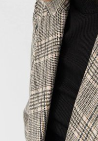 Blazer a cuadros en tonos-beige, negro y marrón, con un cuello estructurado y tejido texturizado, combinado con un cuello de tortuga negro acanalado debajo.