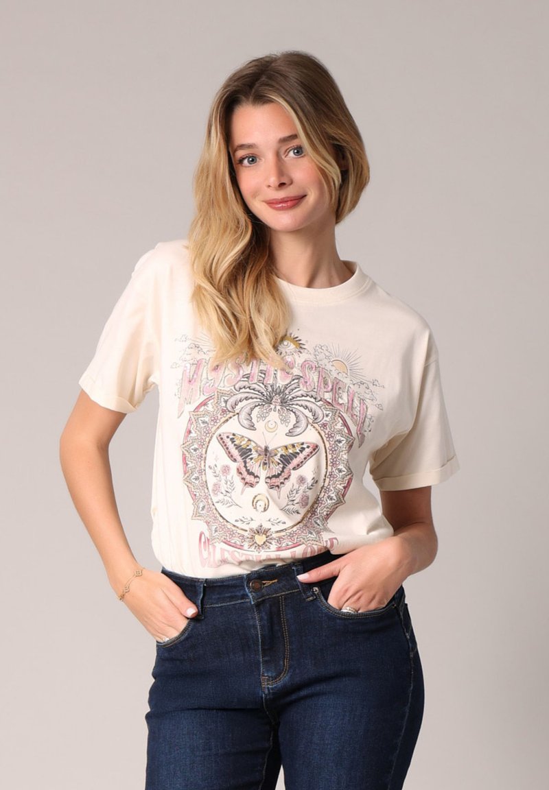 Camiseta crema de manga corta con un gráfico de mariposa colorido y elementos decorativos. Combinada con jeans azul oscuro.