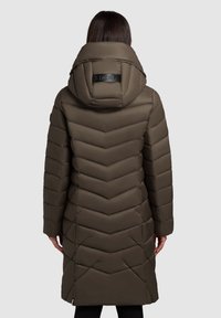 Brūns, garš pūkains mētelis ar ševronu šuvēm, augstu apkakli un logotipa detaļu aizmugurē. Gluda, siltinoša audums, piemērots aukstam laikam.