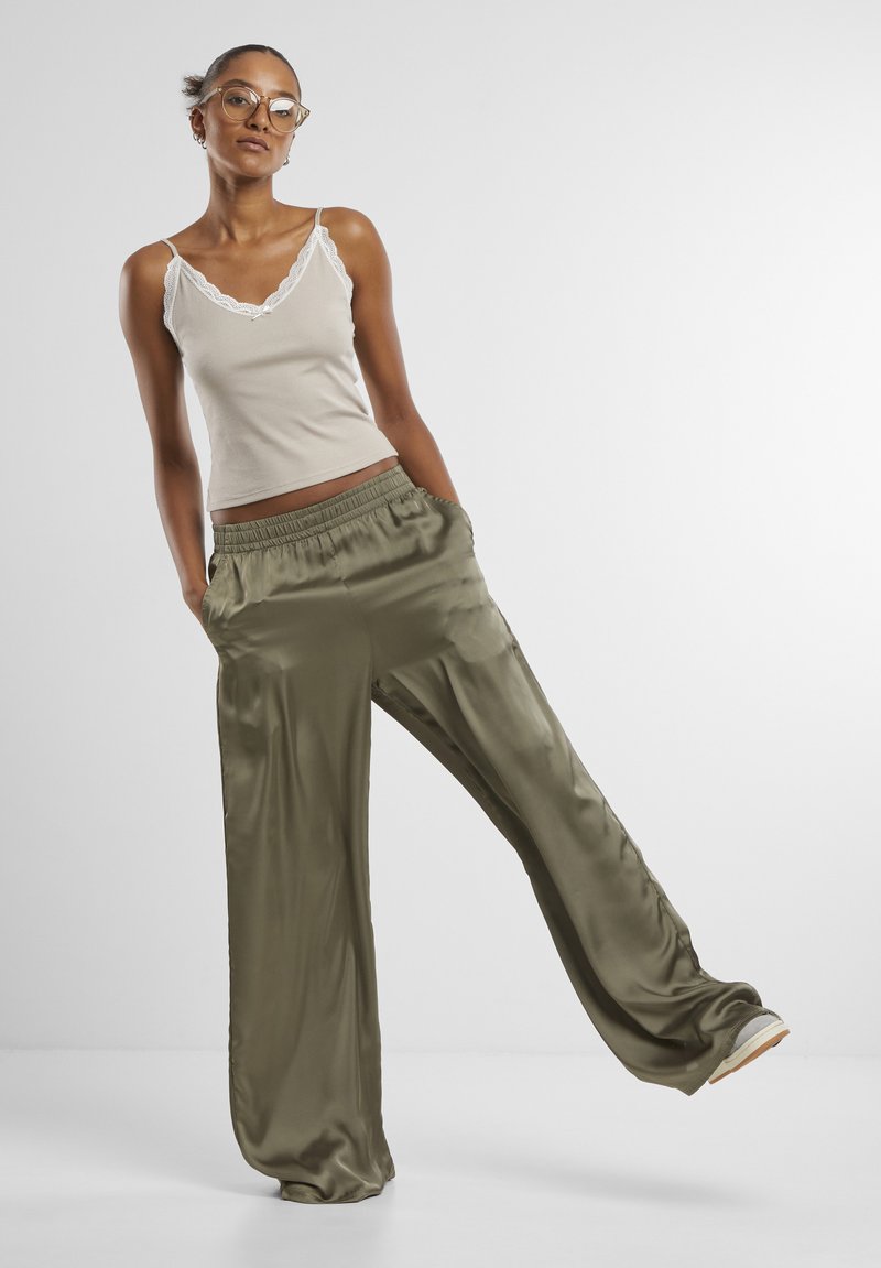 Femme avec lunettes portant un caraco beige orné de dentelle et un pantalon ample en satin vert olive, debout avec une jambe levée sur un fond uni.