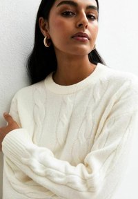 Frau mit dunklen Haaren, die goldene Creolenohrringe und einen weißen Strickpullover trägt, steht mit verschränkten Armen vor einer weißen Wand.