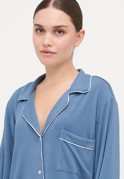Light blue pajama top in soft fabric with a collar, white piping, button placket, and a single chest pocket. Long sleeves.