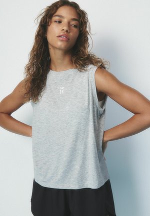 Junge Frau mit lockigem Haar, die ein ärmelloses graues Oberteil mit dem Schriftzug "self" trägt, steht mit den Händen in der Hüfte und schaut seitlich vor einem schlichten Hintergrund.