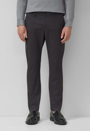 Man draagt zwarte, nauwsluitende broek, grijs shirt met lange mouwen en zwarte leren loafers, staand tegen een effen achtergrond.