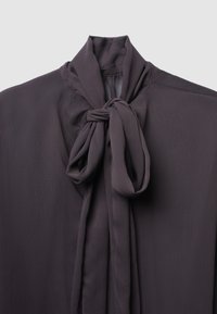 Blusa viola scuro realizzata in un tessuto leggero e texturizzato. Caratterizzata da un colletto annodato drammatico e drappeggi fluente. Design minimalista.