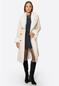 Długi beżowy płaszcz z białym futrzanym kołnierzem i mankietami, złotymi guzikami oraz lekko teksturowanym materiałem, w połączeniu z wysokimi na kolana czarnymi butami.