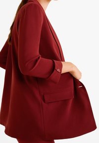 Blazer bordeaux avec une coupe décontractée, en tissu doux, à manches trois-quarts et une poche frontale unique. Texture lisse et design taillé.