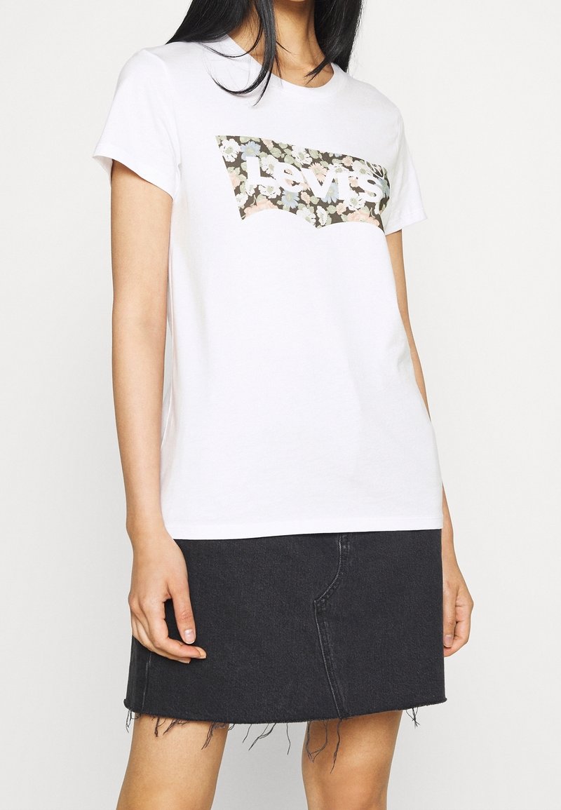 Femme portant un t-shirt Levi's blanc à logo fleuri et une jupe en jean noire à ourlet brut, debout sur fond blanc.