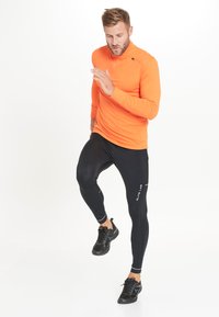 Orange langärmliges Sportshirt kombiniert mit schwarzen Leggings. Das Shirt hat einen hohen Kragen und einen körperbetonten Schnitt, dazu schwarze Sneakers.