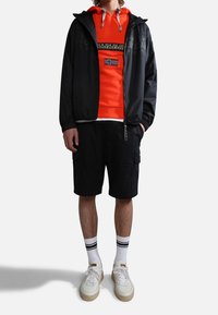 Mężczyzna w pomarańczowej bluzie z kapturem z emblematem flagi, czarnej kurtce zapinanej na zamek, czarnych szortach cargo, białych skarpetach w czarne paski i białych tenisówkach.