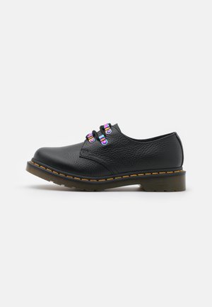 Chaussure basse en cuir noir avec semelle épaisse, coutures jaunes et deux sangles avec boucles iridescentes sur fond blanc.
