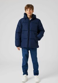 Veste matelassée marine avec capuche, dotée d'une fermeture éclair à l'avant et de poignets élastiques, associée à un jean bleu et des baskets blanches.