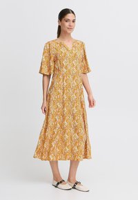 Rochie maxi cu un model paisley galben, cu decolteu în V și mâneci clopot, realizată dintr-un material ușor. Modelul poartă pantofi flats cu accente negre.