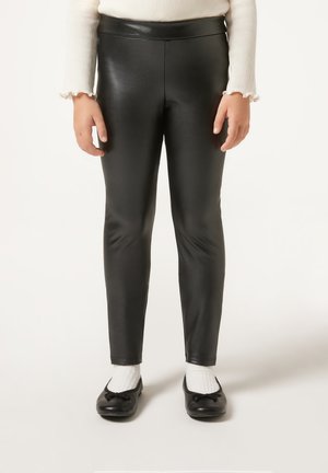 Schwarze, fauxleder Leggings mit glänzendem Finish, schlankem Design und hohem Bund, getragen mit schwarzen Ballerinas und weißen Socken.