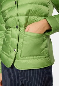 Zielona kurtka puchowa z pikowaną fakturą, wyposażona w boczną kieszeń i zatrzaski. Noszona z granatowymi spodniami w prążki.