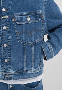 Jeans jakna v svetlo modri barvi, z vodoodbojnimi gumbi, prsno žepom z našito dekoracijo in sproščenim krojem. Teksturirana površina.