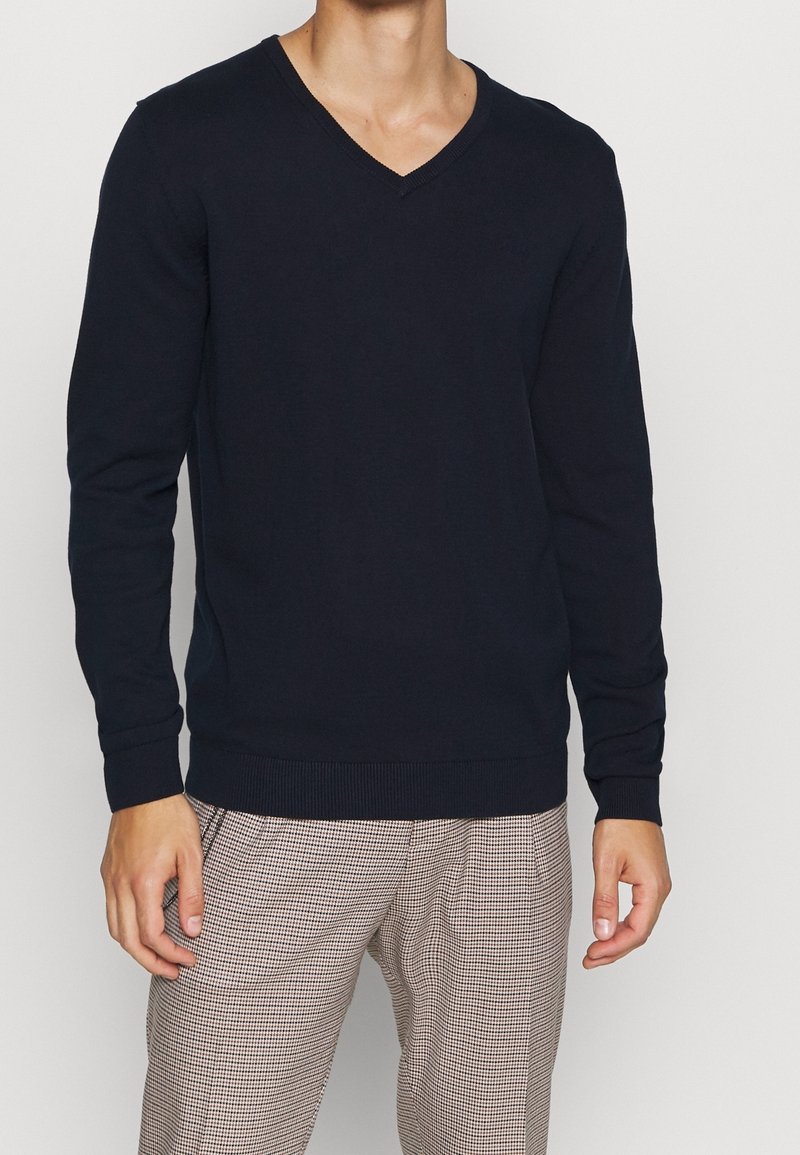 Homme portant un pull bleu marine à col en V et un pantalon beige à carreaux, debout devant un fond blanc.