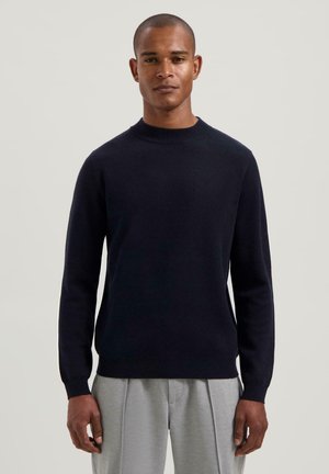 Man wearing a fitted black long-sleeve sweater and light gray pleated trousers standing against a plain light background.