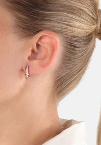 Roséguld hoops örhänge med en böjd design prydd med små klara ädelstenar på ena sidan, visad på en modells öra.