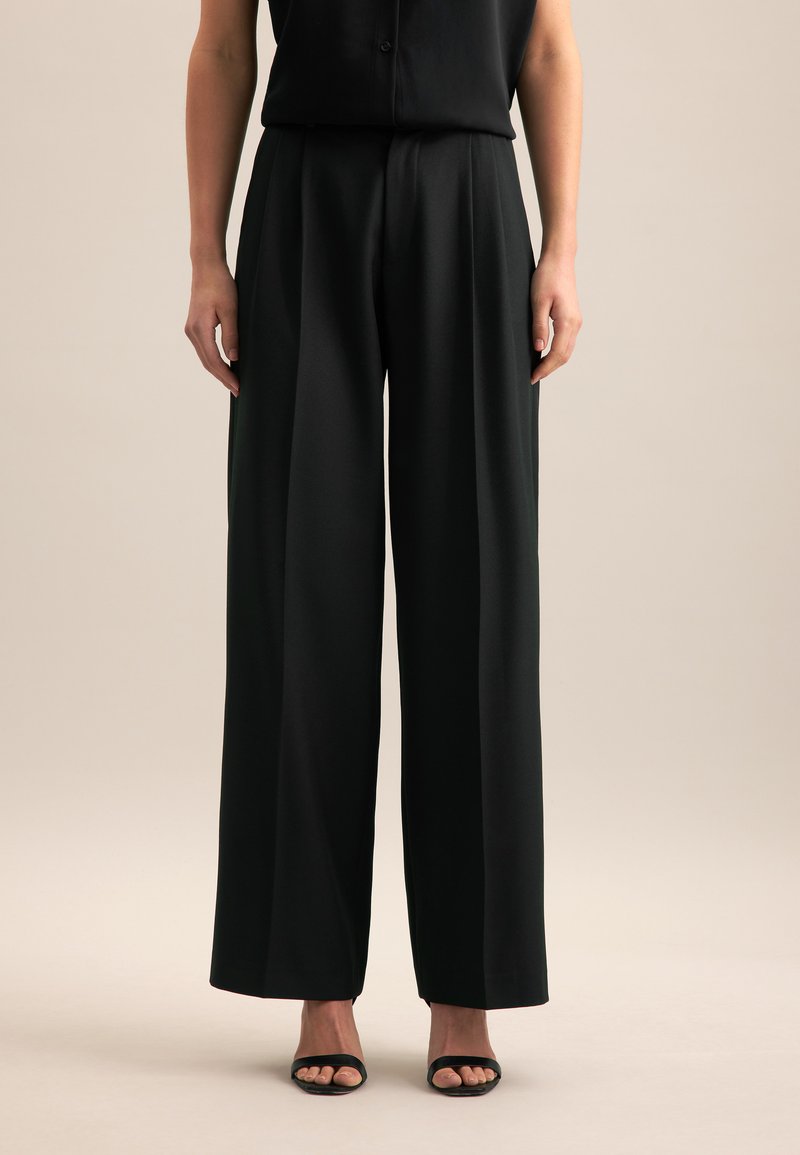 Pantalons larges noirs en tissu lisse, dotés de plis, d'une taille haute et d'une silhouette fluide, associés à des talons noirs.