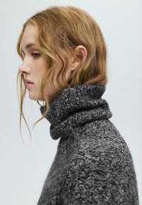 Szürke kötött garbó pulóver texturált mintával, hajtott magas nyakkal és bő szabással, oldalról egy modellen bemutatva.