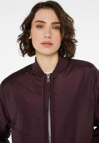 Burgunderfarbene Bomberjacke mit Reißverschluss vorne, geripptem Kragen und glatter Textur. Verfügt über ein minimalistisches Design und eine leichte Konstruktion.