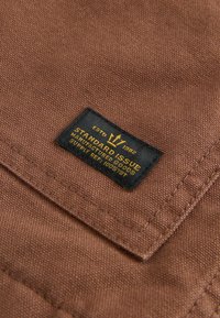 Brunt stof med en struktureret vævning har et sort mærke. Mærket står der "STANDARD ISSUE" på gult, med et krone-logo ovenover teksten.