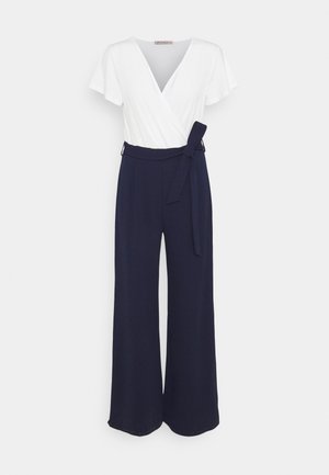 Top bianco in stile avvolgente con maniche corte, abbinato a pantaloni navy a gamba larga con cintura annodata e tessuto liscio.