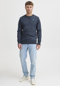 Granatowy sweter z okrągłym dekoltem i ściągaczami, w zestawie z jasnoniebieskimi dżinsami i białymi trampkami. Prosty design, bez wzorów.