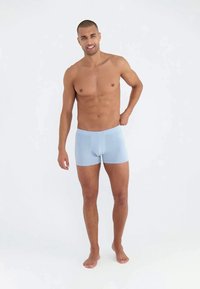 Mosolygó, mezítláb álló férfi világoskék boxeralsót visel egy sima fehér háttér előtt.