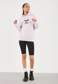 Lys rosa hettegensere med svart grafisk design og tekst, kombinert med svarte knelange shorts og svarte militærstøvler. Hvite sokker synlige.