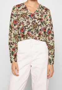 Blommig blus med beige bakgrund, långa ärmar, v-ringning och färgglada blommönster; matchad med ljusa byxor.
