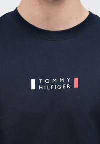 Μπλε ναυτικό t-shirt με στρογγυλή λαιμόκοψη, κατασκευασμένο από βαμβάκι, που διαθέτει λευκό και κόκκινο λογότυπο γραφικού "TOMMY HILFIGER" τοποθετημένο στο κέντρο του στήθους.