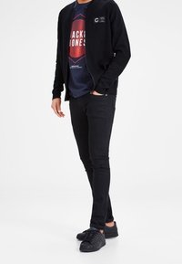 Svart zip-up jacka, mörk grafisk t-shirt med röd och blå mönster, svarta jeans och svarta sneakers. Enkla, avslappnade kläder med modern design.