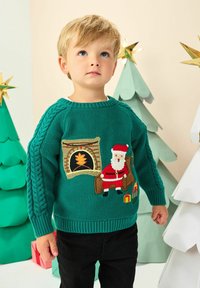 Junger Junge in grünem Weihnachts-Pullover mit Santa, Kamin und Geschenke-Design, der zwischen dekorativen Papier-Weihnachtsbäumen steht.