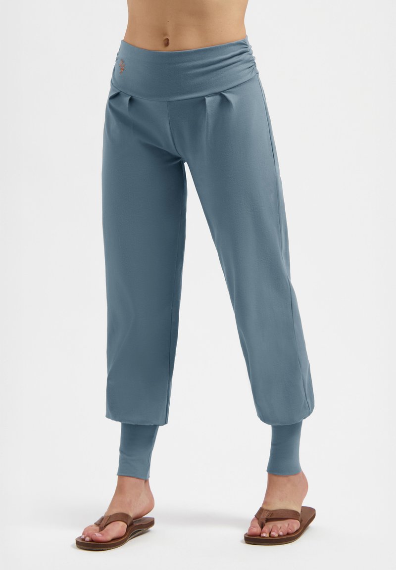 Blaue, weit geschnittene, verkürzte Hose mit elastischen Bündchen, getragen mit braunen Flip-Flops, die den Bauch und die Unterschenkel vor einem weißen Hintergrund zeigen.