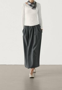 Jupe longue grise avec taille froncée, associée à une chemise blanche à manches longues et une écharpe à carreaux noir et blanc. On porte des ballerines de couleur claire.