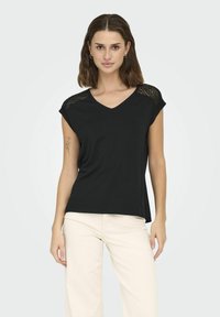 Czarny T-shirt z krótkimi rękawami, dekoltem w serek i koronkowymi wykończeniami na ramionach. Miękka tkanina o luźnym kroju. Model połączony z beżowymi spodniami.