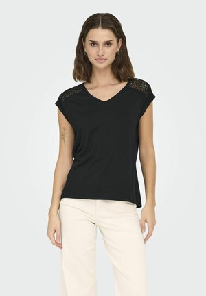 Czarny T-shirt z krótkimi rękawami, dekoltem w serek i koronkowymi wykończeniami na ramionach. Miękka tkanina o luźnym kroju. Model połączony z beżowymi spodniami.