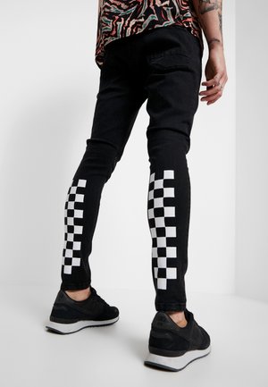 Osoba ubrana w czarne dżinsy z białymi wzorami w kratę na łydkach, czarne tenisówki, stojąca na jednolitym tle.