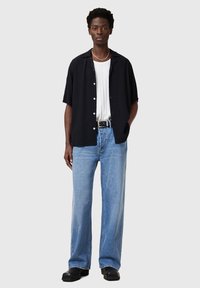 Schwarzes kurzärmliges Hemd mit strukturiertem Muster, weißes geripptes Tanktop, hellblaue weit geschnittene Jeans und schwarze klobige Schuhe. Silberne Schmuckakzente.