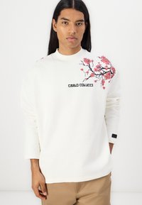 Sweat-shirt blanc à manches longues, avec une broderie florale rose sur une épaule et le logo "CARLO COLUCCI" en noir. Texture lisse.