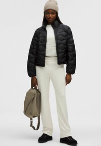 Mujer con un gorro beige, chaqueta negra acolchada, parte superior y pantalones blancos, sosteniendo una mochila topo, y usando zapatos negros sobre un fondo blanco.