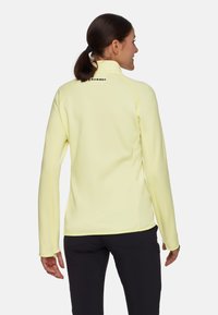 Giacca in pile gialla con collo alto, design aderente e cuciture piatte. Presenta un logo sulla parte posteriore del collo e maniche lunghe con fori per i pollici.