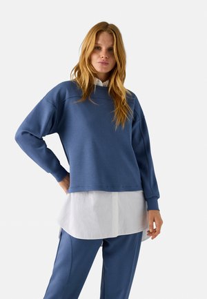 Sudadera azul con un corte holgado, mangas largas, puños acanalados, sobre una camisa blanca con cuello, combinada con pantalones azul a juego.