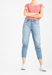 Persona de pie con los brazos cruzados, usando una camiseta de manga corta rosa, unos jeans recortados de color azul claro y zapatillas blancas, sobre un fondo blanco.