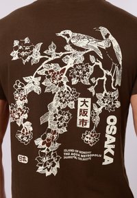 Branche fleurie avec deux oiseaux perchés au-dessus d'un texte japonais, des coordonnées et « Osaka » sur un fond en tissu marron.