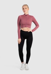 Geribbelde, cropped zip-up top in mauve met lange mouwen, gecombineerd met hoge zwarte leggings en witte sport schoenen.