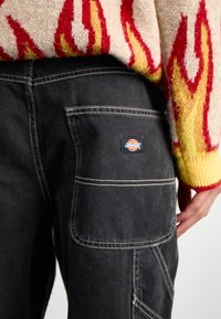 Črni jeans hlače z velikim stranskim žepom, belim šivanjem in značko. Nad njimi je viden pulover z večbarvnim vzorcem ognja.