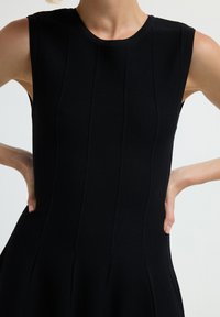 Abito nero senza maniche caratterizzato da un tessuto testurizzato, corpetto aderente con cuciture verticali e gonna svasata. Collo liscio e forma strutturata.